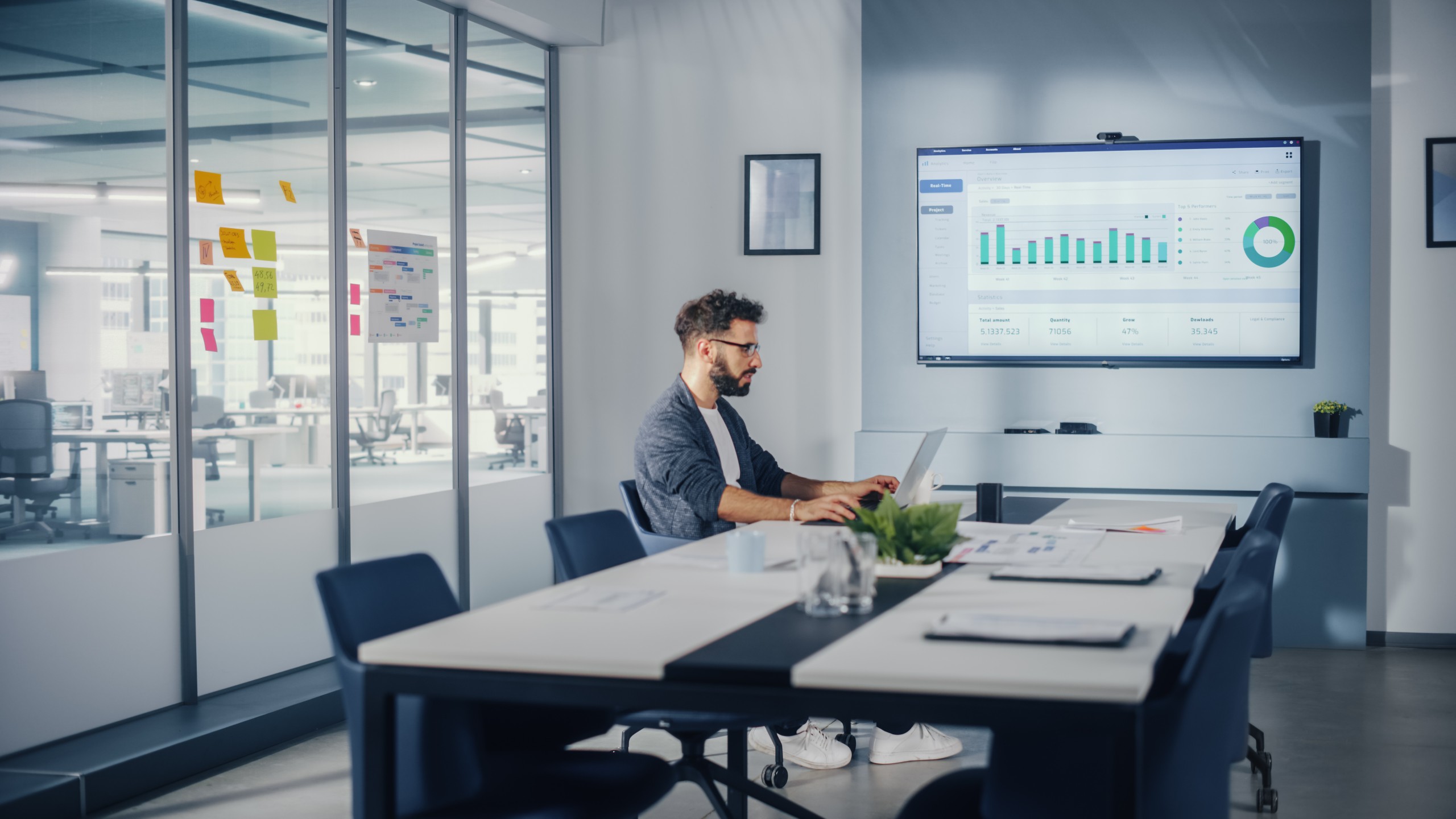 Office Conference Meeting Room: Handsome Hispanic Specialist Working on Laptop Computer with Big Wall TV with Big Data Analysis, Charts and Infographics. Businessperson Working in Growing Startup.